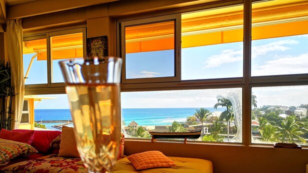 A room with a bed, a window with a view of the ocean, and a hand holding a glass.
