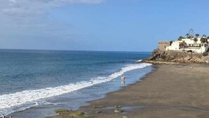 A beach with a building on a cliff, a clear sky, and calm sea.