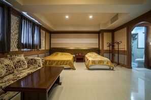Quarto de hotel com duas camas, um sofá, uma mesa de madeira e um banheiro visível através de um arco.