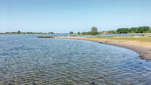 A calm lake with clear water, a sandy beach, and a distant shoreline with trees and buildings.