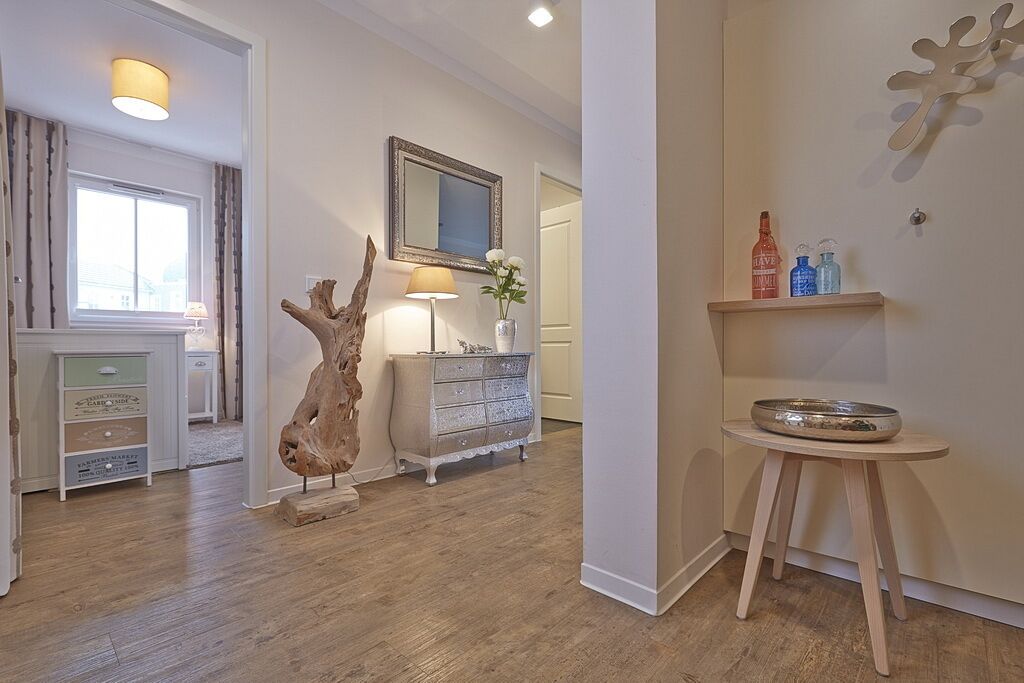 A room with a wooden floor, a dresser with drawers, a mirror, a vase with flowers, and a shelf with bottles.