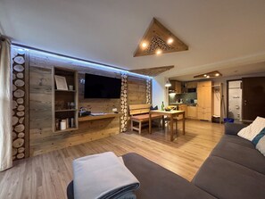 Ein gemütliches Wohnzimmer mit einem Flachbildfernseher, Holzdekor, einem Esstisch mit Stühlen und einem Sofa.