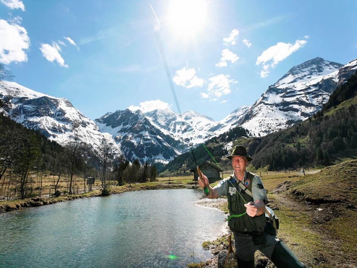 Eine Person angelt an einem Bergsee, im Hintergrund schneebedeckte Gipfel.