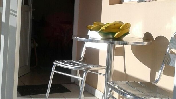 A small patio area with a table and two chairs, a bowl of bananas on the table, and a window with blinds. - Stunning villa top 10mins beach (Rivière-Pilote)