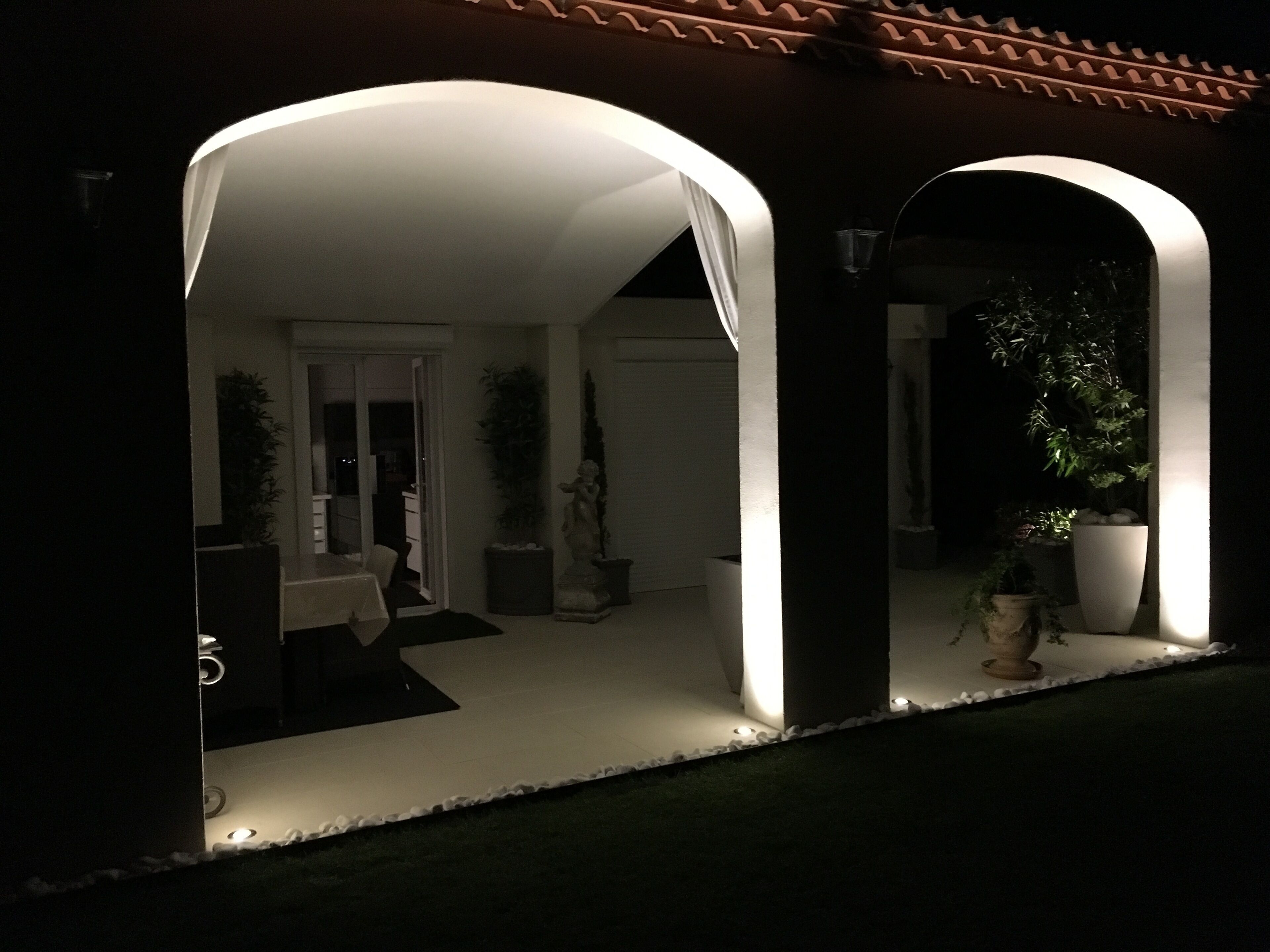 A modern outdoor patio area with a white ceiling and columns, surrounded by potted plants and a statue.