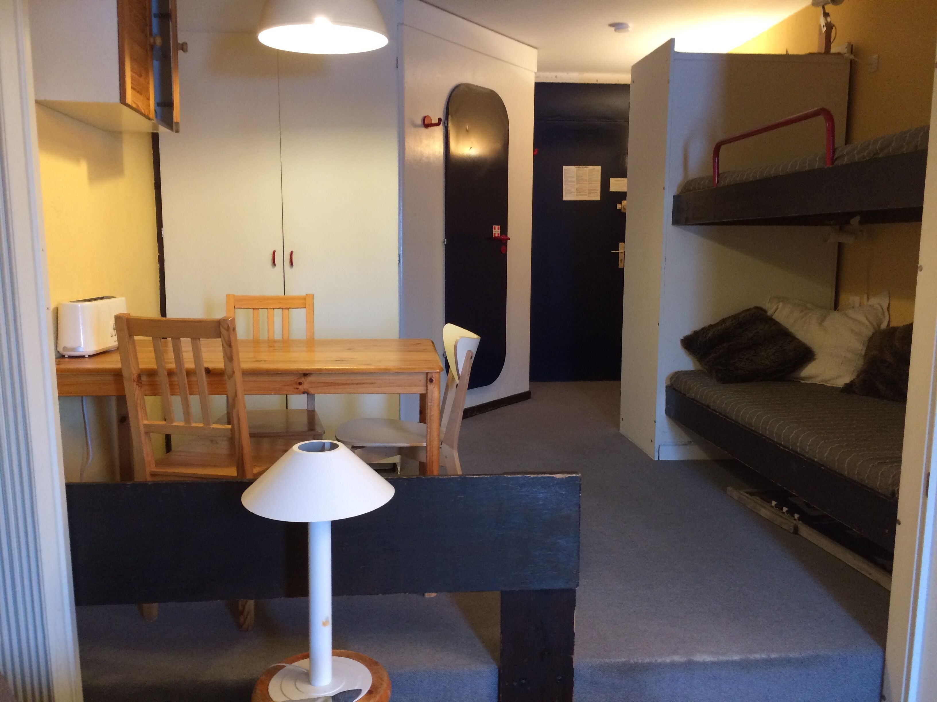Une pièce de taille modeste comprenant une table à manger en bois, des chaises, un banc, un lit superposé et une lampe.