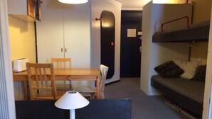 Een kleine kamer met een houten eettafel, stoelen, een bankje, een stapelbed en een lamp.