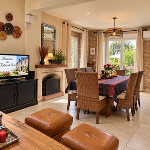 Un comedor con sillas de mimbre, una mesa cubierta con mantel, chimenea y televisor mostrando un mensaje de bienvenida.
