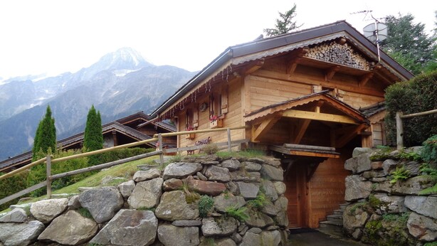 Eine Holzhütte in den Bergen mit Steineingang, Holztreppe und Blick auf schneebedeckte Gipfel.