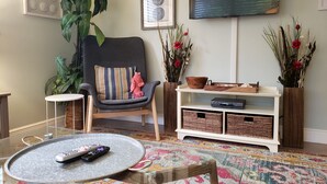 A living room with a grey armchair, a round coffee table, a flat-screen TV, and a bookshelf with wicker baskets. - The Little Sandy...#1 in Siesta! (Siesta Key)
