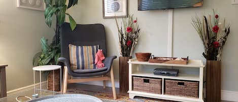 A living room with a grey armchair, a round coffee table, a flat-screen TV, and a bookshelf with wicker baskets.