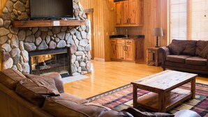 A cozy living room with a stone fireplace, wooden cabinets, a leather sofa, and a television mounted on a stone wall. - Private - Secluded Lake Home/Sleeps up to 20 people (Deerwood)