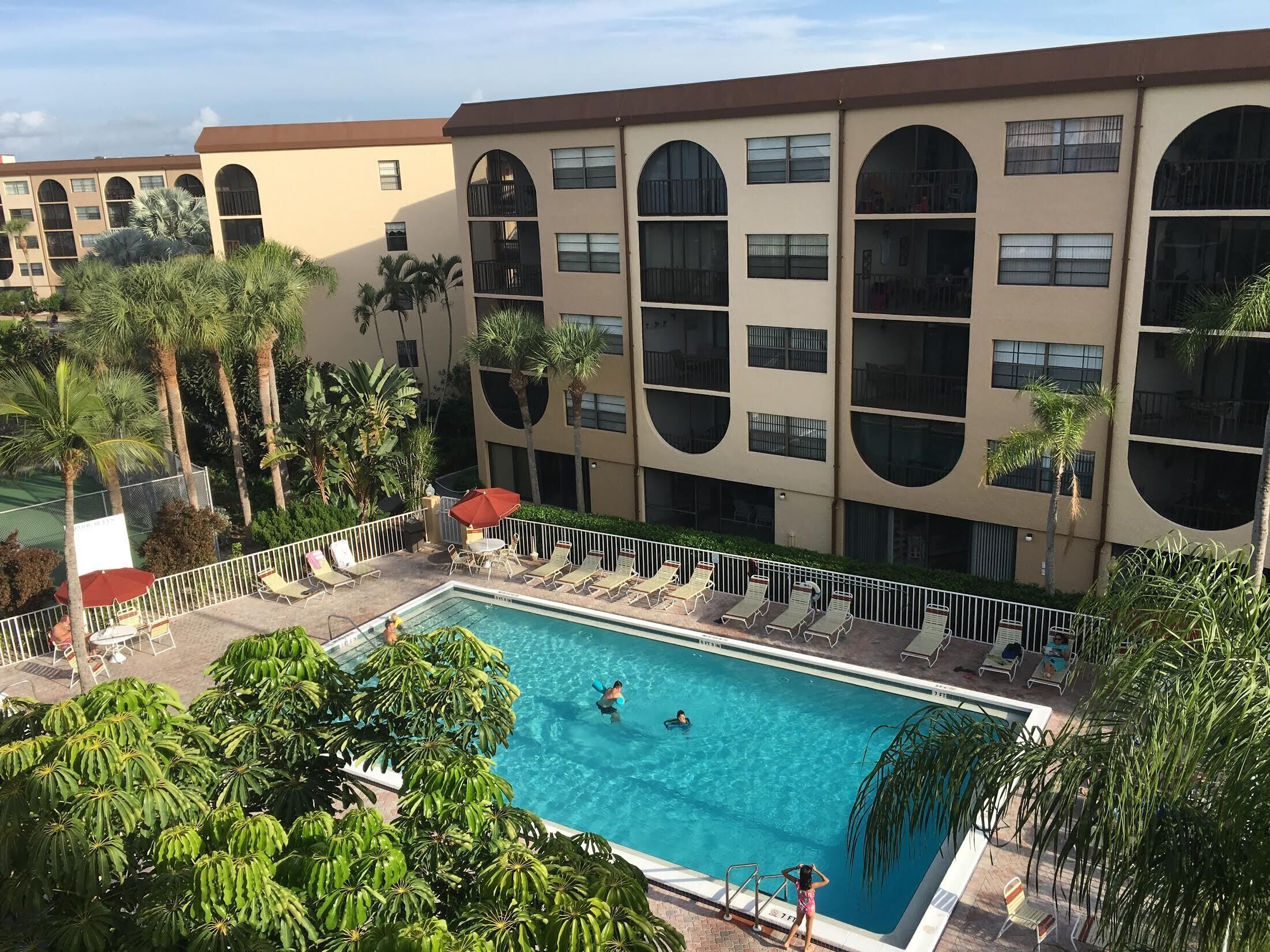 A swimming pool with lounge chairs and palm trees, surrounded by a multi-story building with large windows and balconies.