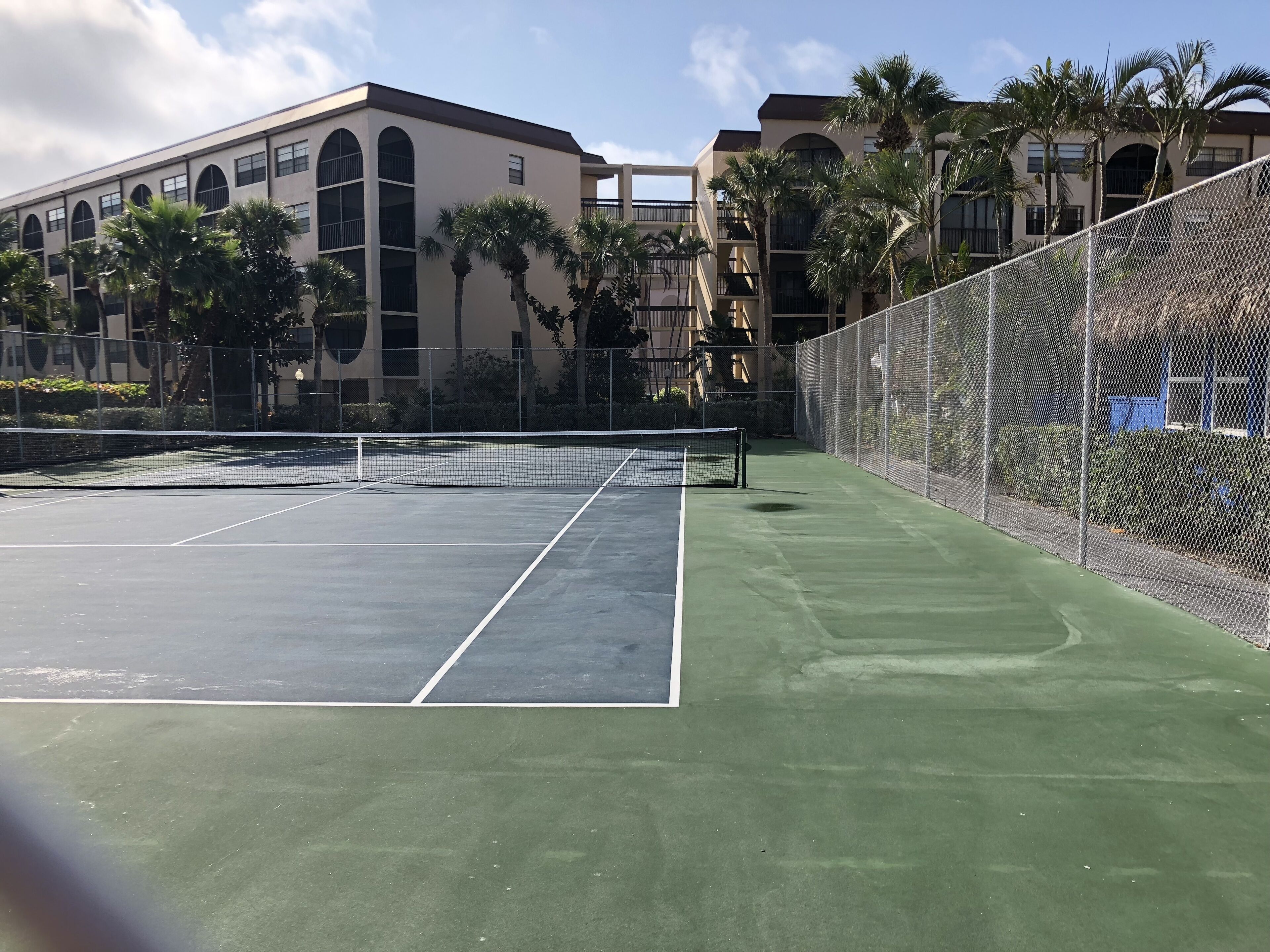 An outdoor tennis court with a net, surrounded by a chain-link fence and palm trees.