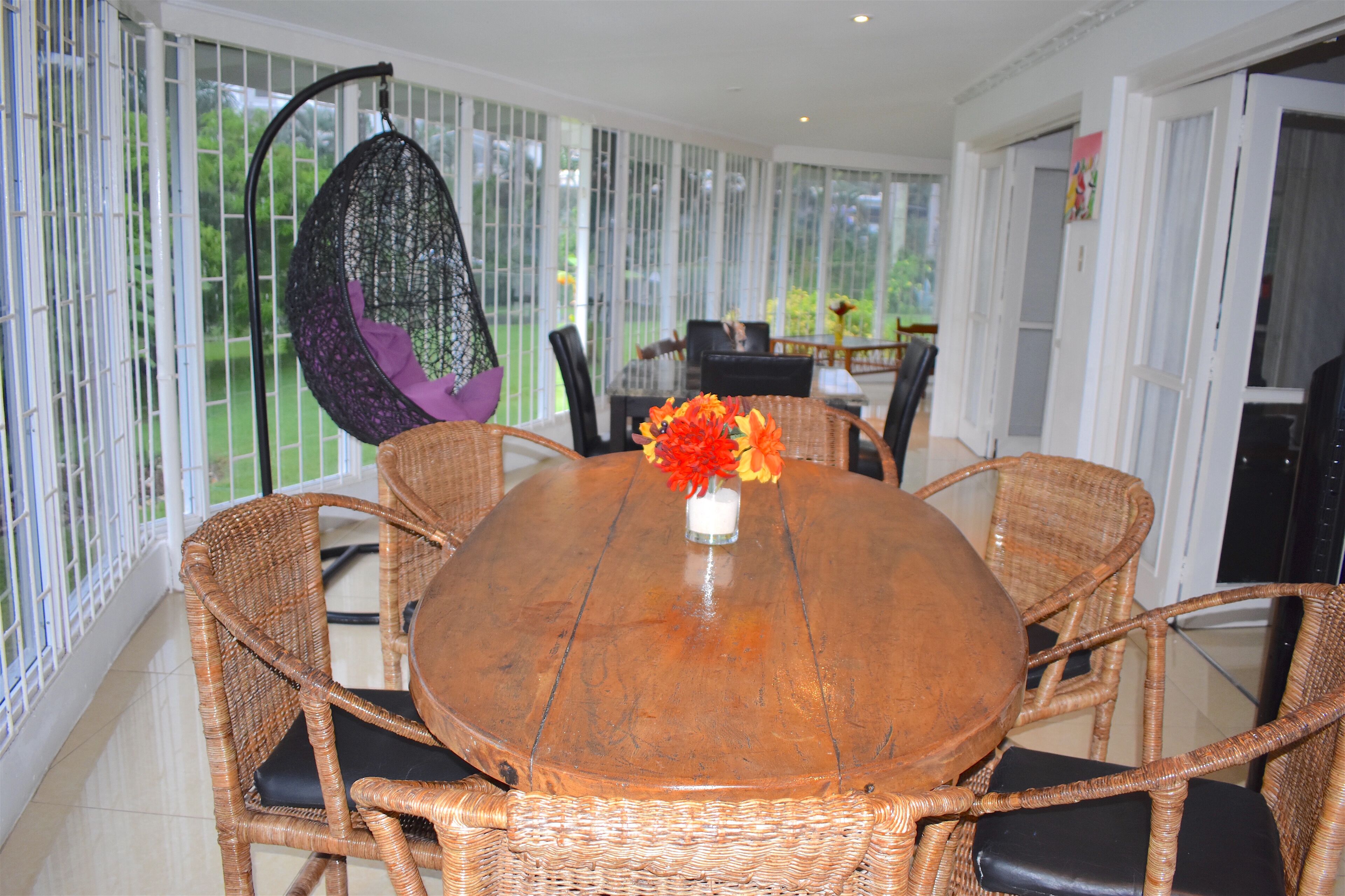 A round wooden table with wicker chairs, a hanging chair, and a vase with flowers.