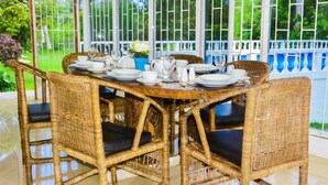 Une table à manger dressée avec de l’assiette blanche, des tasses et des couverts, entourée de chaises en osier avec des coussins noirs, dans une pièce dotée de grandes fenêtres et offrant une vue sur la verdure.