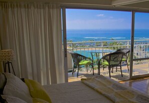 Quarto de hotel com uma grande porta de vidro deslizante que dá para uma varanda com vista para o oceano e a praia. Há duas cadeiras de vime na varanda.