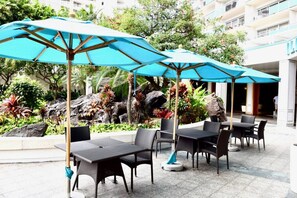 Mobiliário de exterior com guarda-sóis, um jardim ajardinado e um edifício ao fundo.