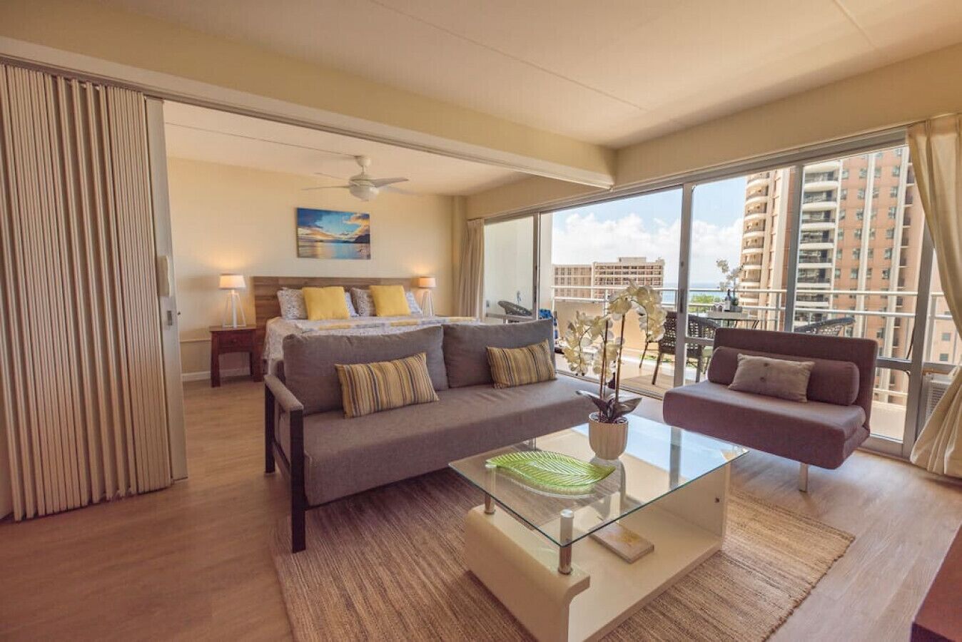 A modern hotel room with a large bed, a sofa, a glass coffee table, and a view of the city through large windows.
