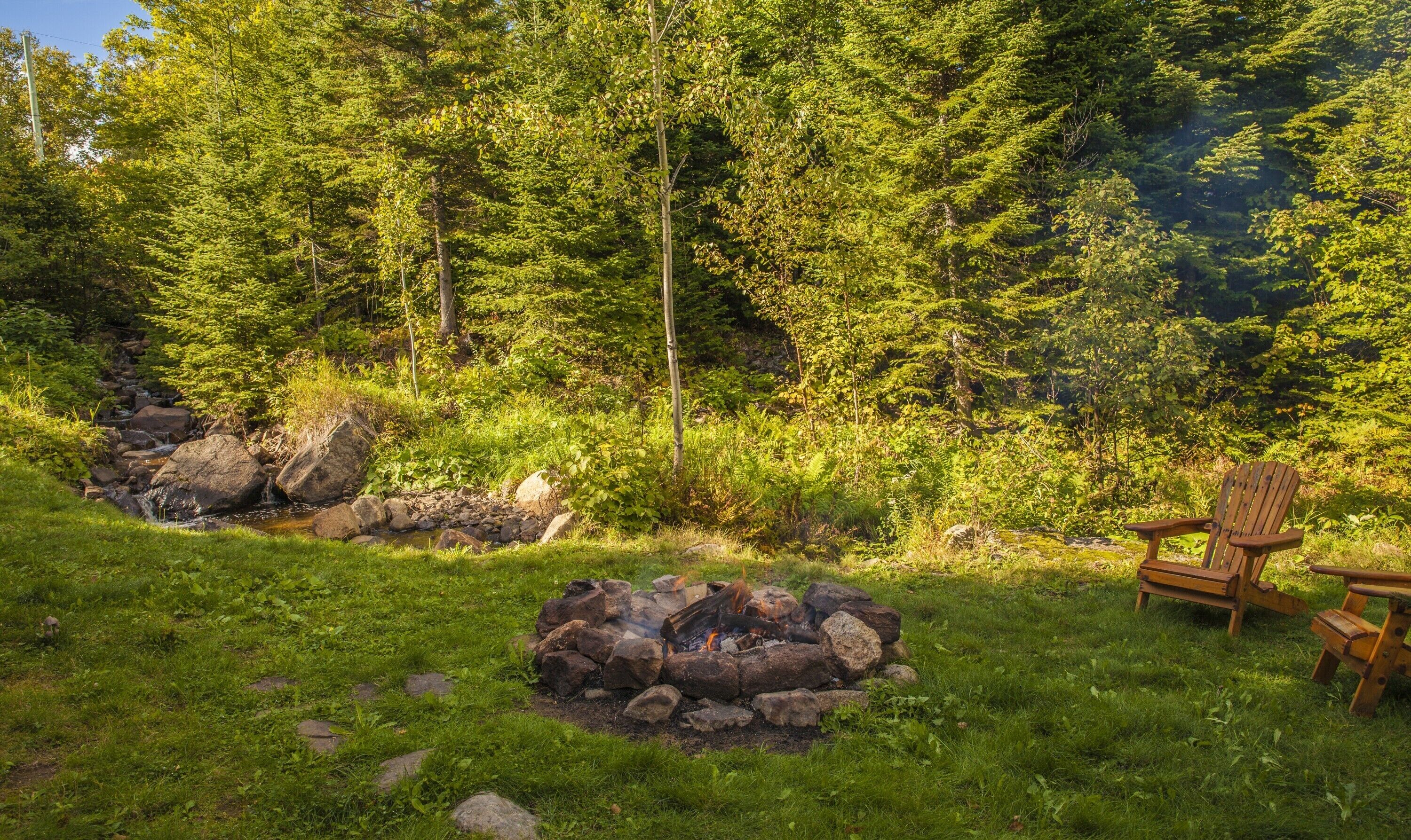 Um camping com lareira, uma cadeira e um riacho em área arborizada.
