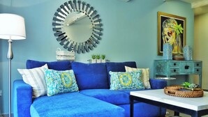 Un sofà blau amb coixins estampats, un mirall rodó, un quadre i una taula petita amb objectes decoratius.