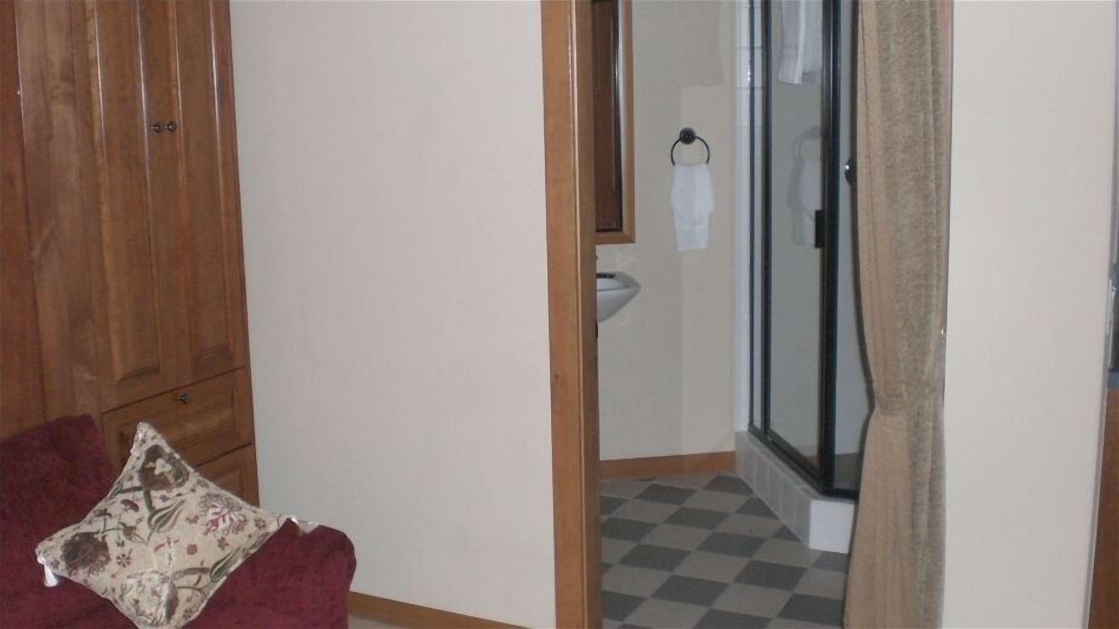 A room with a wooden wardrobe, a checkered floor, a shower area with a glass door, and a red sofa with a patterned pillow.