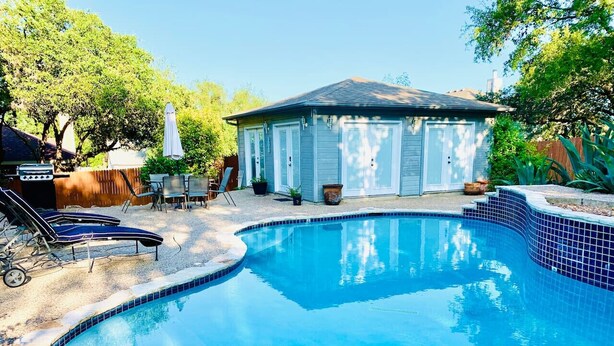 A swimming pool with a curved edge, a covered patio area, and a small building in the background.