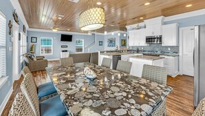A spacious dining area with a large table surrounded by wicker chairs, a kitchen with white cabinets and stainless steel appliances, and a ceiling with wooden beams and recessed lighting. - Amazing View, Gorgeous Beachfront, Huge Deck, Luxury Retreat-Sleeps 14, Starlink (Crystal Beach)
