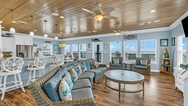A spacious living area with a large sectional sofa, wicker furniture, and a round coffee table. There are ceiling fans, pendant lights, and a view of the ocean through large windows. - Amazing View, Gorgeous Beachfront, Huge Deck, Luxury Retreat-Sleeps 14, Starlink (Crystal Beach)