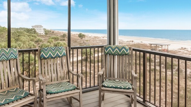 A covered outdoor seating area with wooden chairs and green patterned cushions, overlooking a beach with a distant building.