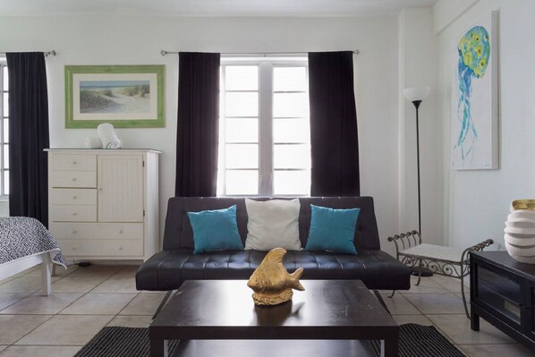 Een woonkamer met een zwarte bank, twee blauwe kussens, een houten salontafel met een sculptuur van een vis, een wit kastje, een bed met een beddegoed met een patroon, en een schilderij aan de muur.