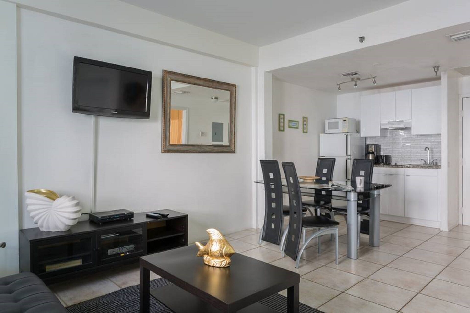 A modern living room with a flat-screen TV, a dining area with a glass table and chairs, and a kitchen with white cabinets and a microwave.