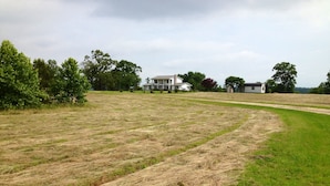 A rural landscape with a large open field, a white house with a porch, and a few trees in the background. - Fully Furnished Two Bedroom Farm House, "The Little House" Has Everything ! (Delight)