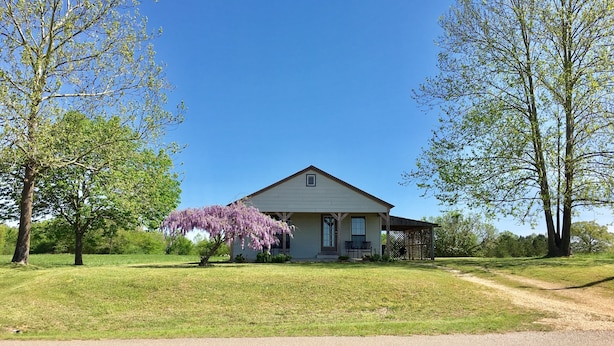 A single-story house with a front porch, surrounded by a grassy lawn and trees. - Fully Furnished Two Bedroom Farm House, "The Little House" Has Everything ! (Delight)