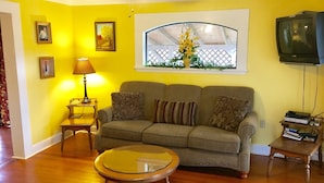 A living room with a sofa, a glass coffee table, a television, and a window with a view of a fence and flowers. - Fully Furnished Two Bedroom Farm House, "The Little House" Has Everything ! (Delight)