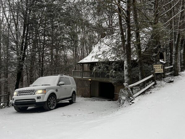 Un bosque nevado con una camioneta SUV blanca estacionada cerca de una cabaña.
