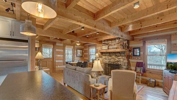 A log cabin interior with a stone fireplace, wooden beams, and a cozy seating area. - Cozy Log Cabin, Mountain views, HOT TUB, & Fire Pit- accepting renters! (Zionville)