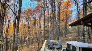 A deck with outdoor seating and a grill area, surrounded by a forest with autumn foliage. - Cozy Log Cabin, Mountain views, HOT TUB, & Fire Pit- accepting renters! (Zionville)