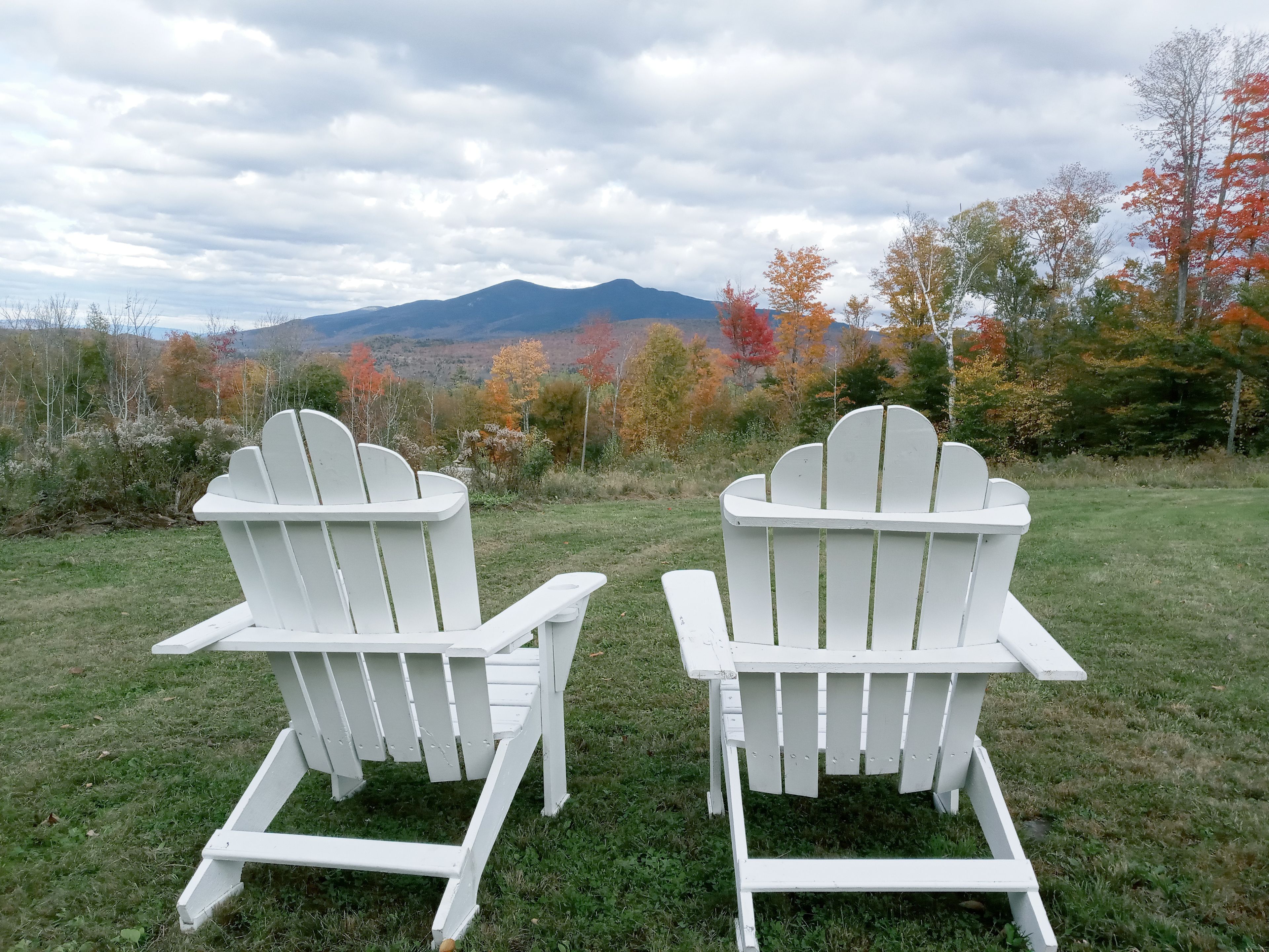 To hvide Adirondack-stole med udsigt til et bjergrigt landskab med efterårsfarver.