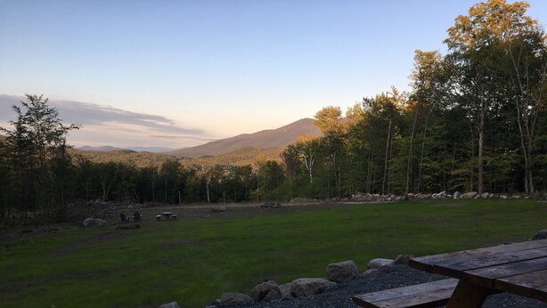 A picnic area with a table and benches, surrounded by a grassy field and trees, with mountains in the background.