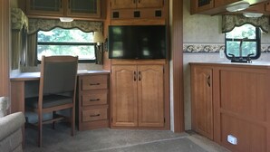A compact RV kitchen with wooden cabinets, a built-in TV, a dining area with a chair, and a window with curtains.