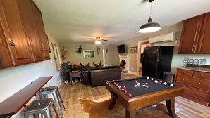 A pool table, bar stools, a sofa, and a fireplace in a living room with wooden floors and a ceiling fan.