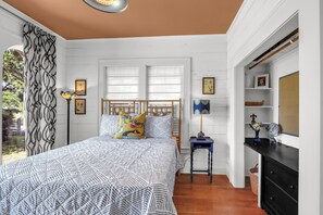 A bedroom with a bed, a nightstand, a dresser, a window with blinds, and a door leading to another room.