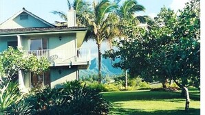 Une maison dotée d’un balcon, entourée d’une végétation luxuriante et de palmiers.