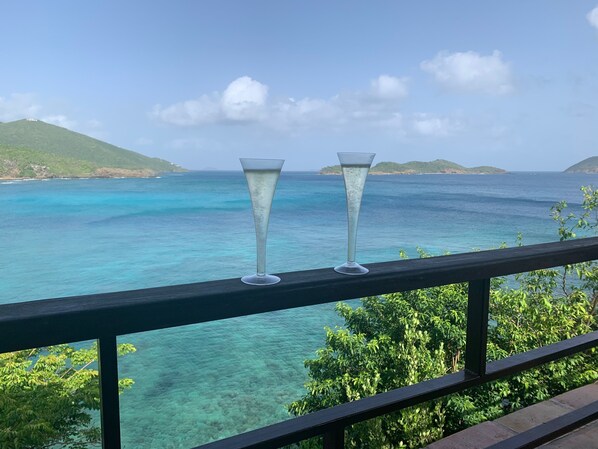 Two champagne flutes on a balcony overlooking a turquoise sea with distant green hills.