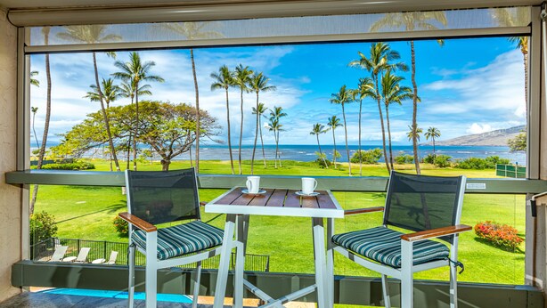 A balcony with two chairs and a table, overlooking a grassy area with palm trees and a view of the ocean. - Leinaala Oceanfront #302 Panoramic Ocean Front View, A/C Great Location, Pool (Kihei)