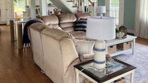 A spacious living room with a high ceiling, a staircase, a large beige sofa, a glass-top coffee table, and a view of the outdoors.