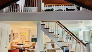 A two-story house with a wooden staircase, a dining area with a chandelier, and a balcony with a railing.