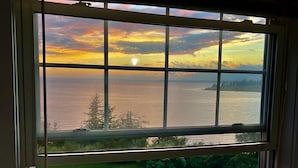 A view through a window of a grassy outdoor area with a table and chairs, overlooking a body of water and a distant shoreline.