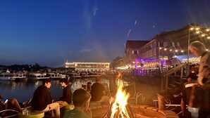 Ein Außenbereich am Flussufer mit Sitzgelegenheiten und einem Feuerkorb, mit Blick auf einen Fluss und ein beleuchtetes Gebäude im Hintergrund.